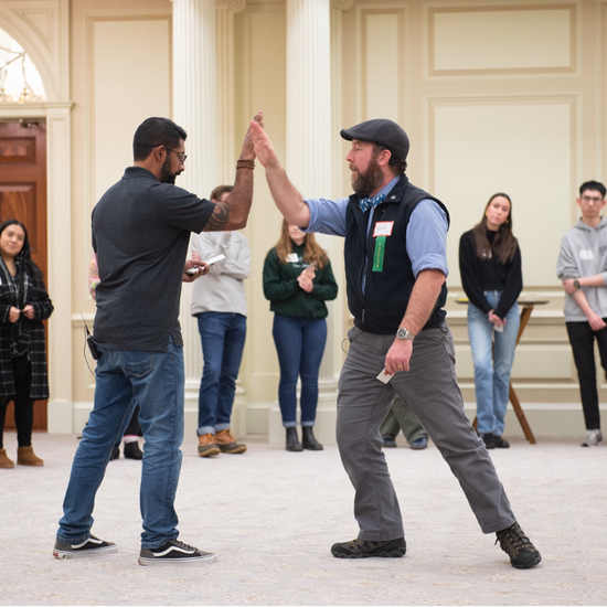Two faculty members are seen giving each other a high-five, surrounded by a large circle of students.
