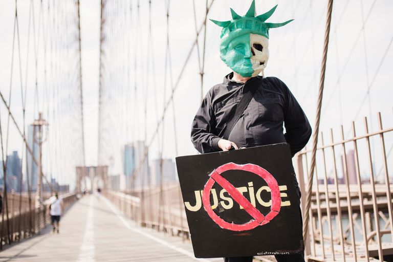Individual on a bridge holding a sign with "Justice" crossed out, wearing a unique mask.